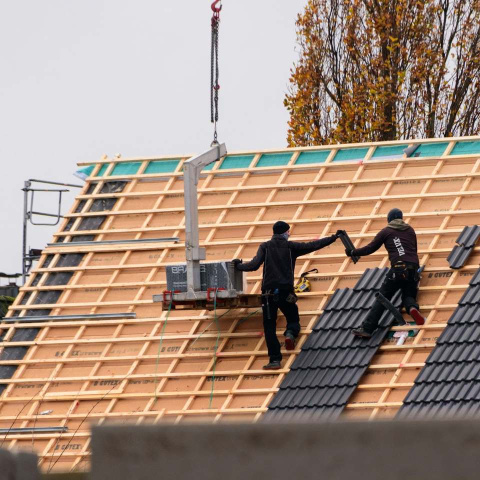 Two roofers laying dark tiles on new battens, with materials lifted up by crane